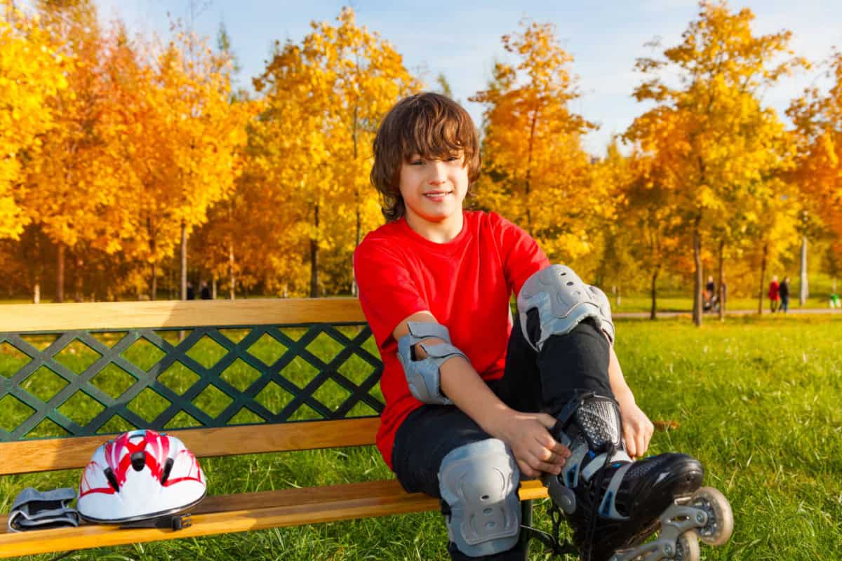 Un băiat cu echipament de protecție își pune patinele cu rotile pe o bancă din parc