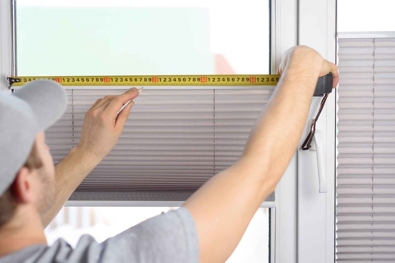 O persoană măsoară lățimea unei ferestre cu o ruletă și marchează poziția cu un creion pentru montarea unei jaluzele plisate.