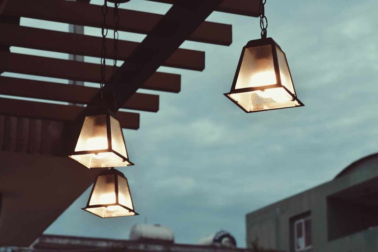 Trei felinare de exterior iluminate cu rame metalice și geamuri texturate care atârnă de o pergolă din lemn pe fundalul cerului în amurg.