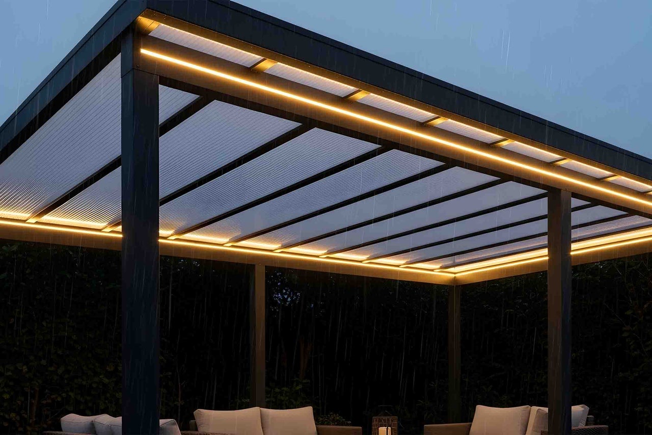 Acoperiș de terasă iluminat, din metal, cu acoperiș translucid și benzi LED alb cald deasupra unei zone de relaxare în aer liber pe ploaie.