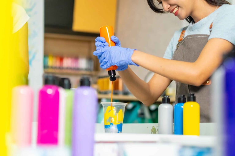 Femeie cu mănuși turnând vopsea dintr-o sticlă într-o cană; sticle de vopsea în prim-plan