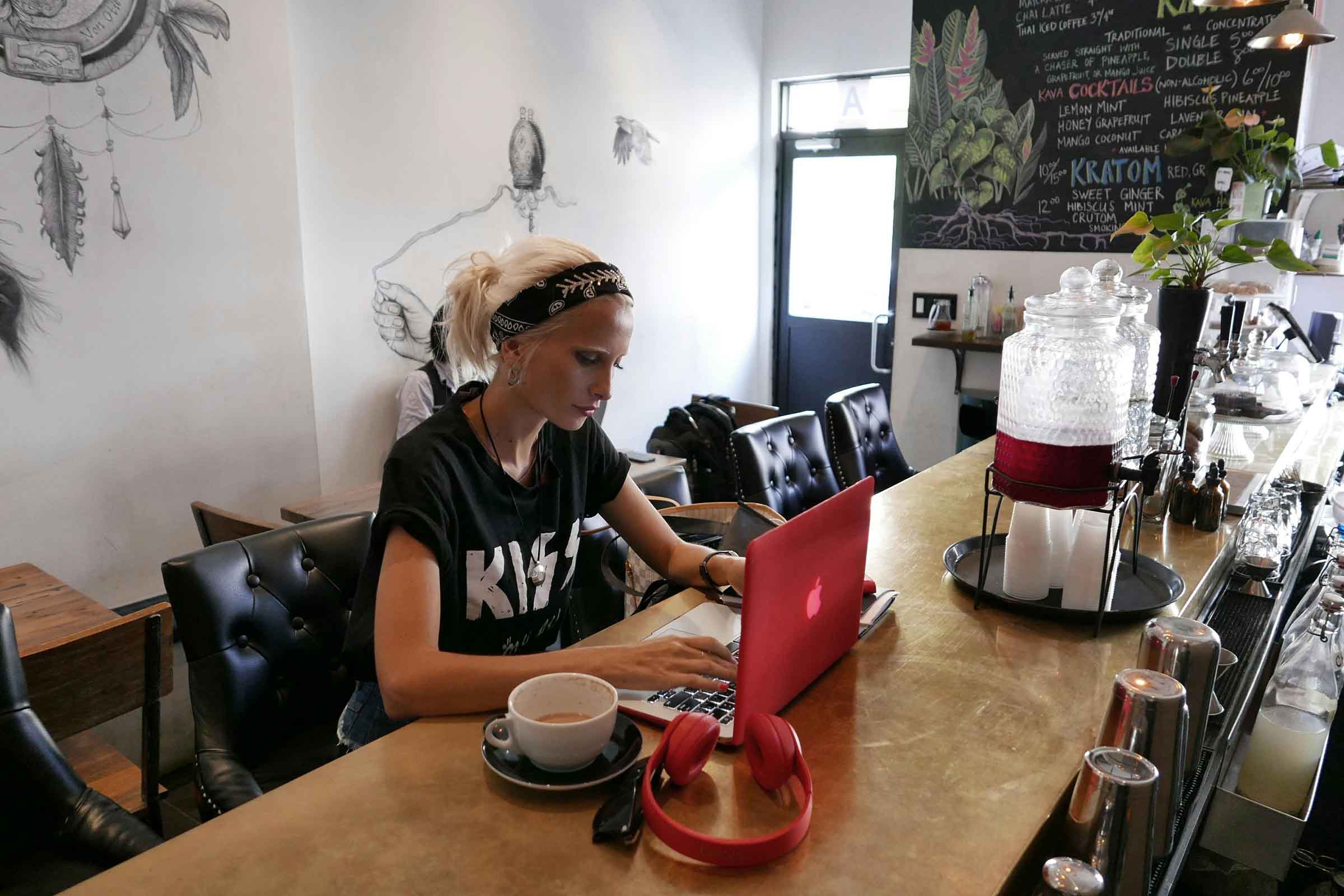 Scenă într-o cafenea cu o femeie lucrând la un laptop, o ceașcă de cafea și căști pe masă.