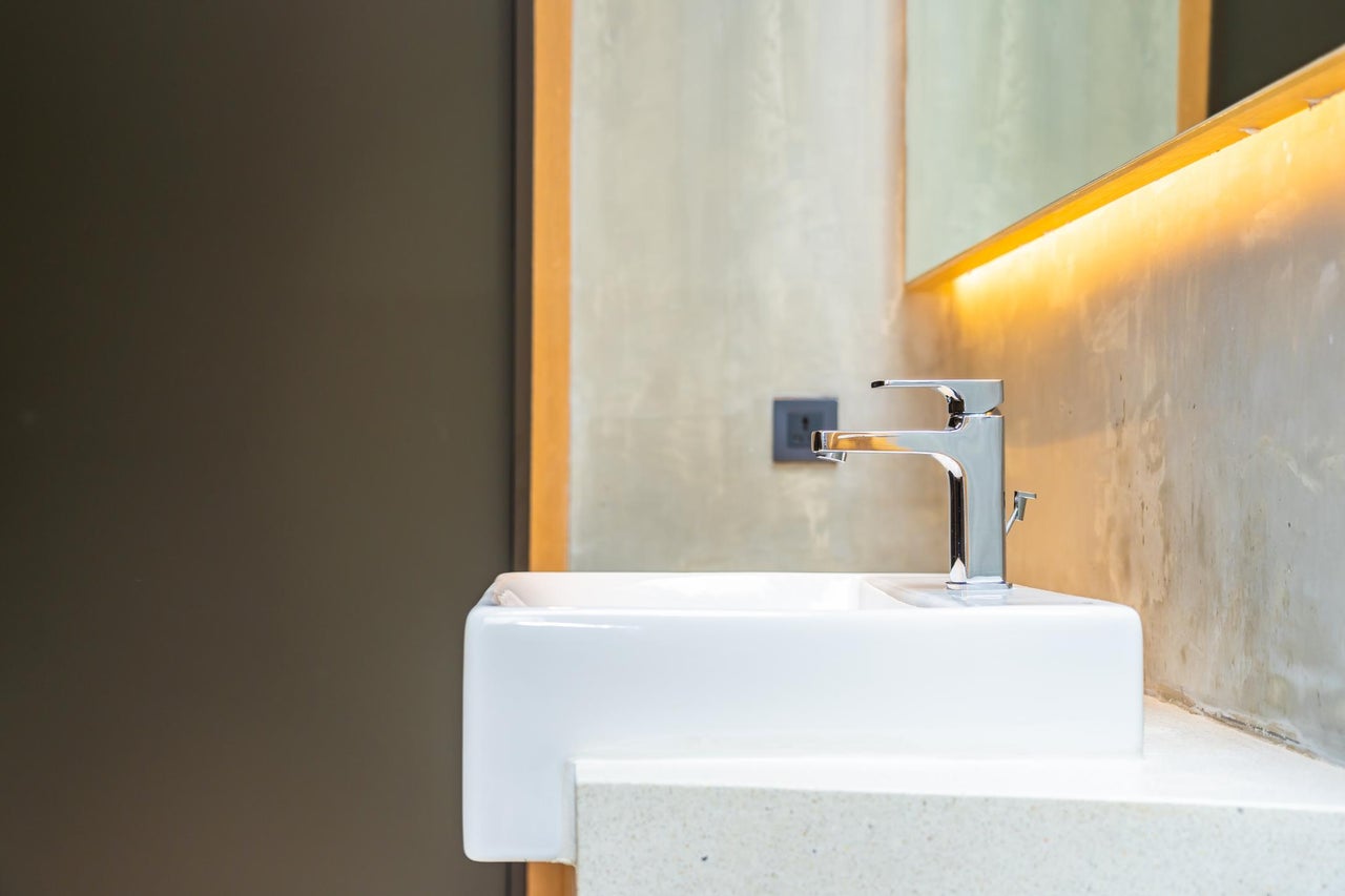 Lavoar modern din ceramică albă cu baterie monocomandă cromată într-o baie luminoasă cu perete din beton și oglindă cu iluminare LED.