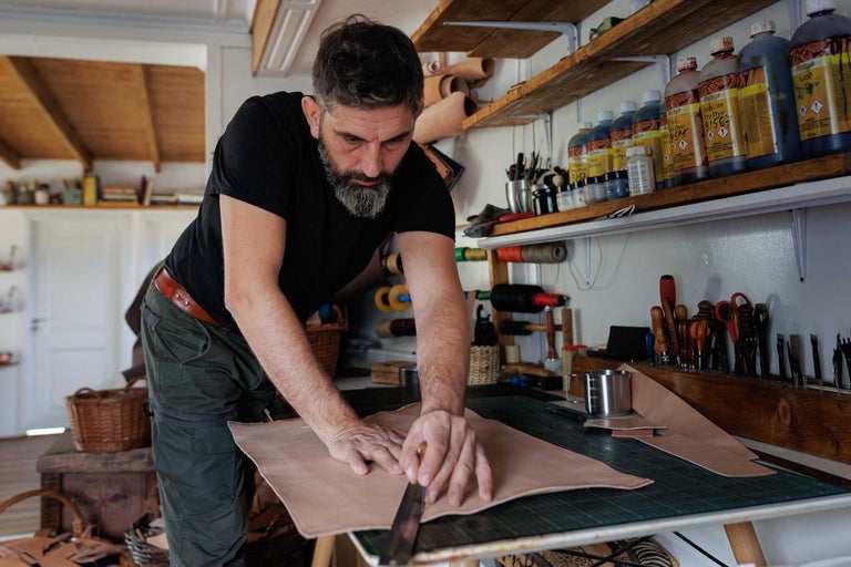 Un meșter taie piele pe o masă într-un atelier. Rafturi cu unelte și vopsele de piele sunt vizibile în fundal.