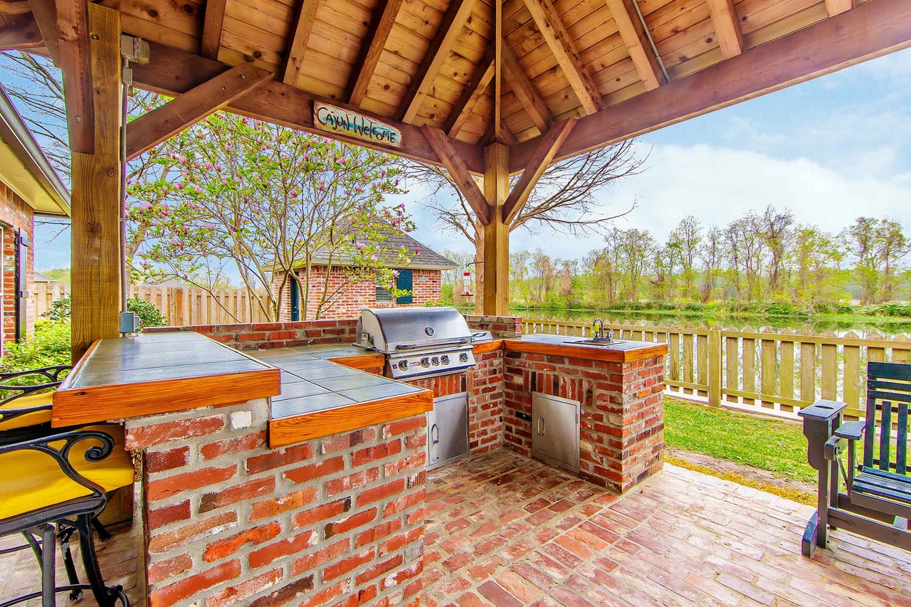 Bucătărie exterioară sub un acoperiș de lemn cu grătar, chiuvetă și blaturi de lucru din cărămizi.