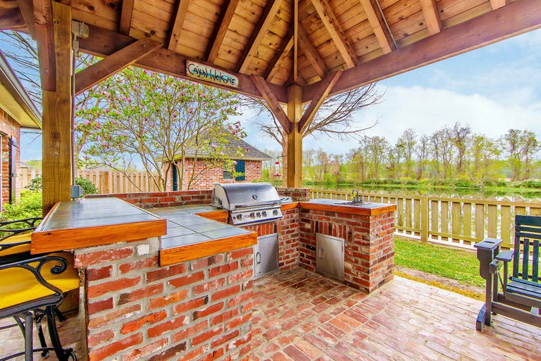 Bucătărie exterioară sub un acoperiș de lemn cu grătar, chiuvetă și blaturi de lucru din cărămizi.