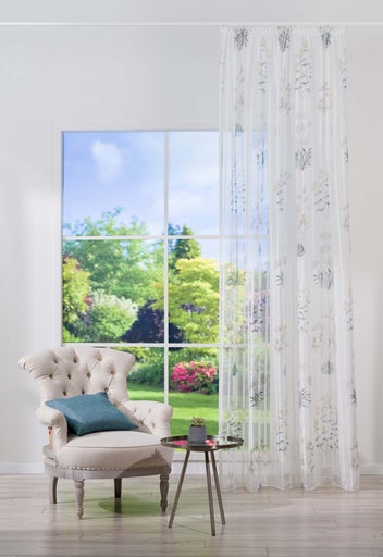 Cameră de zi cu fotoliu, măsuță laterală și perdea transparentă cu model floral în fața unei ferestre cu vedere spre grădină