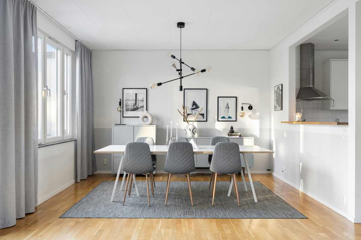 Sufragerie modernă luminoasă cu masă albă, scaune tapițate gri, covor gri, candelabru negru și pardoseală din lemn.