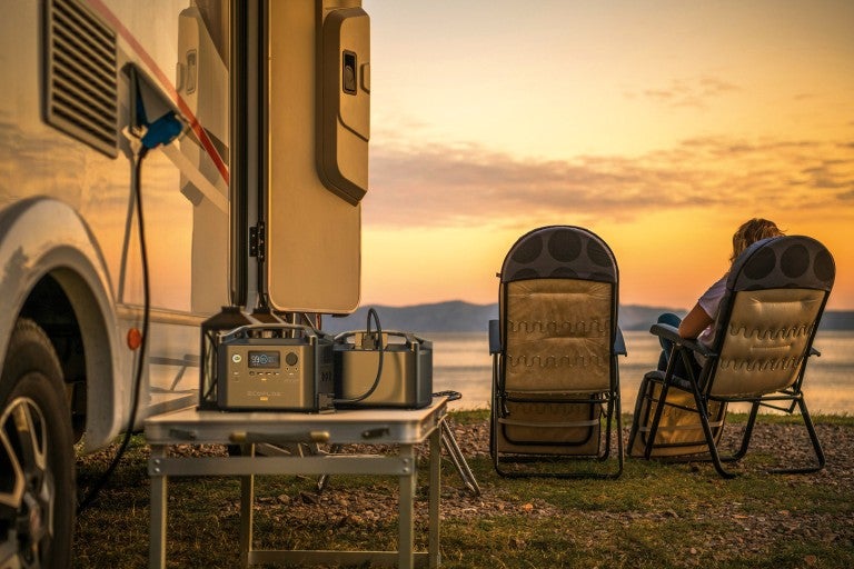 Scenă cu o rulotă, centrală electrică portabilă pe o masă și două scaune de camping la apus
