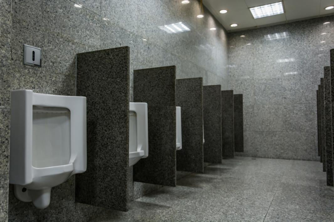 Rând de pisoare albe din ceramică într-o cameră sanitară luminoasă, placată cu gresie, cu panouri despărțitoare din granit.