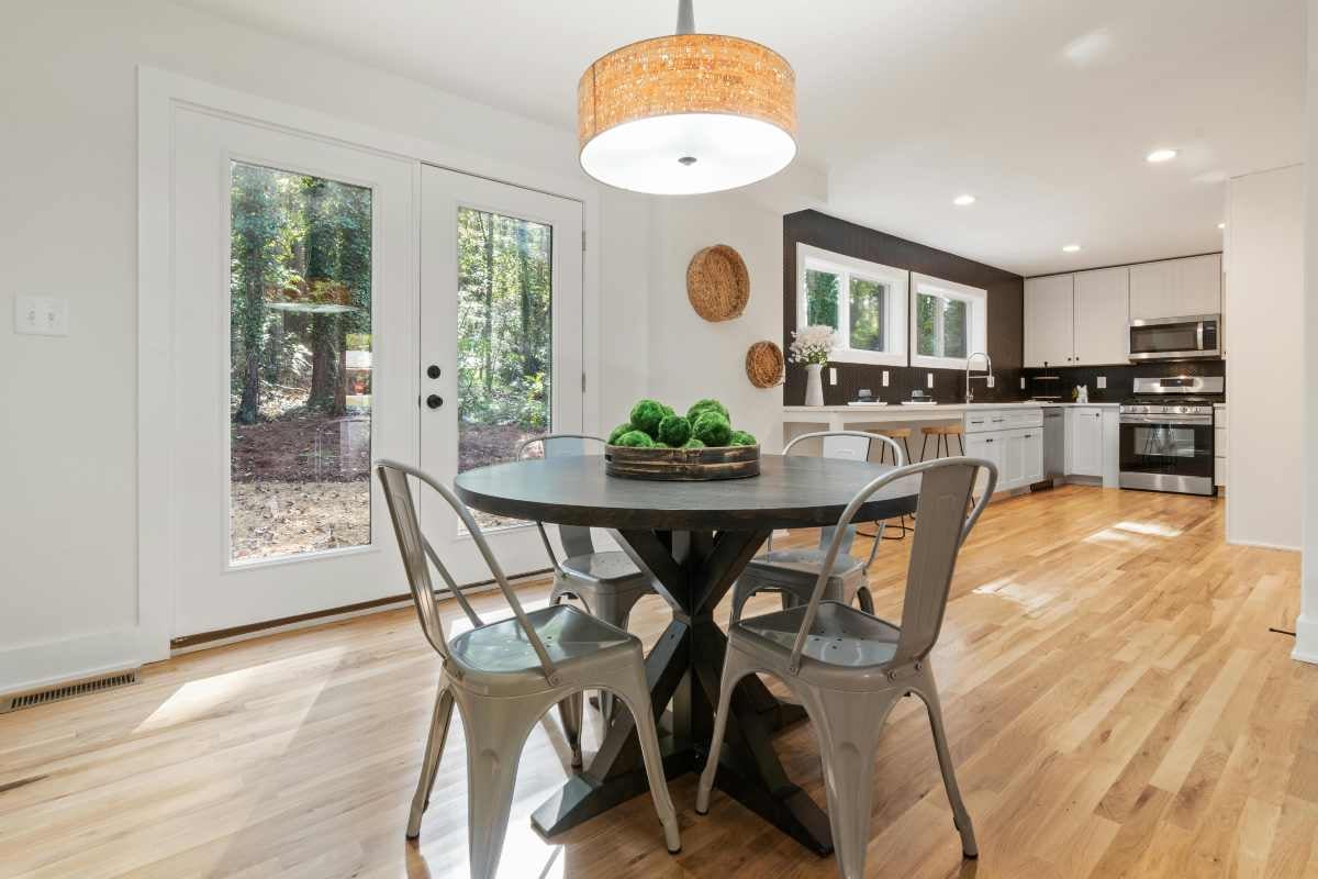 Zonă de luat masa luminoasă cu masă rotundă din lemn, scaune metalice, lustră și bucătărie modernă în fundal cu podea din lemn.