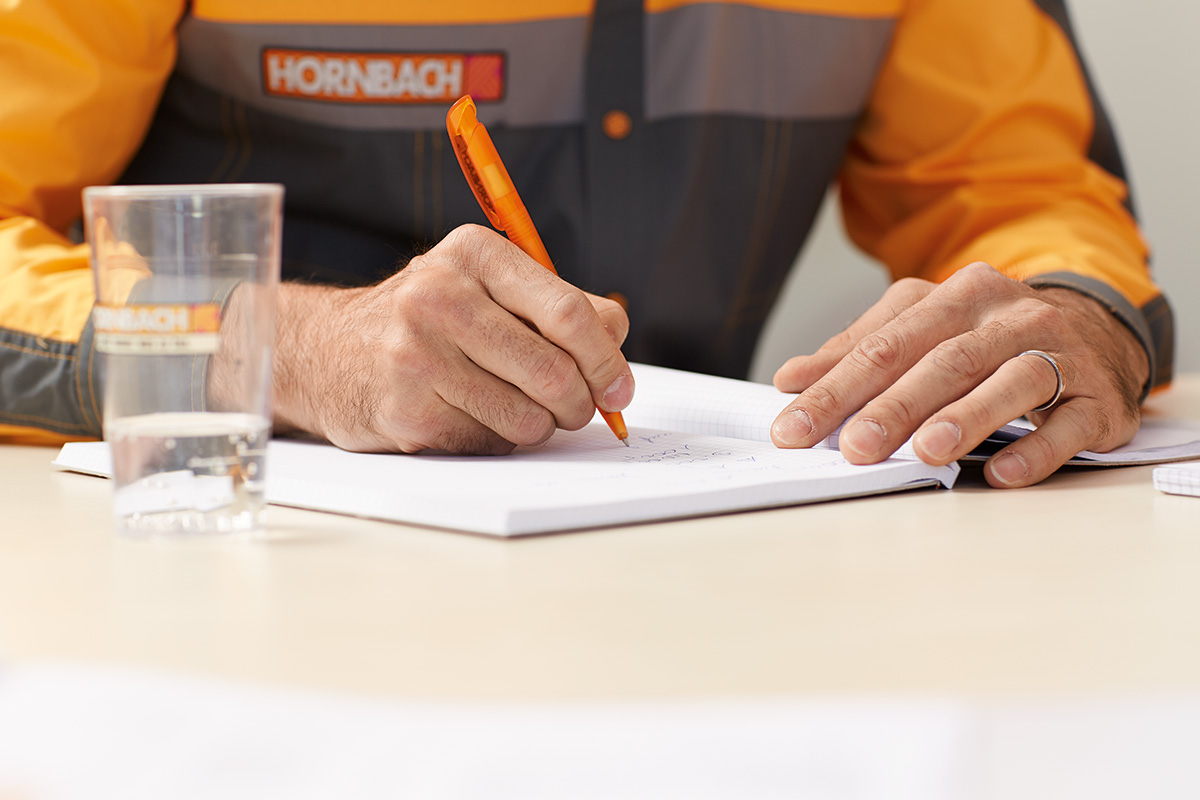 O persoană în ținută de lucru Hornbach scrie într-un carnet cu un stilou.