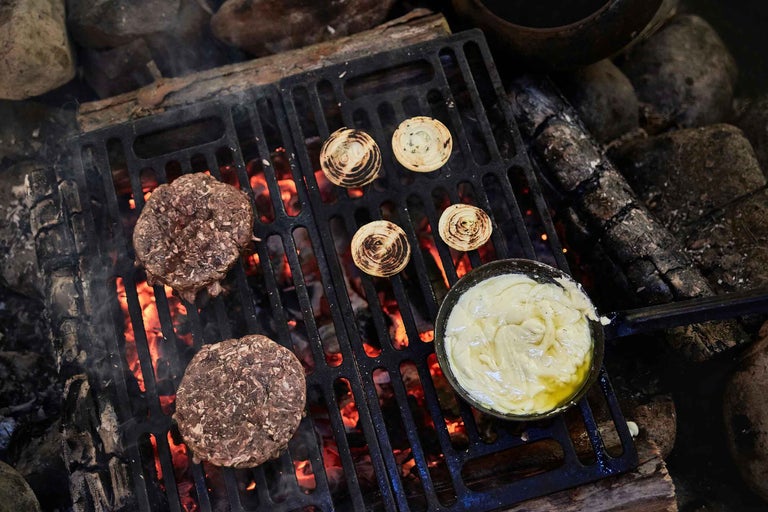 Două burgeri, rondele de ceapă și un castron cu sos pe un grătar deasupra unui foc deschis.