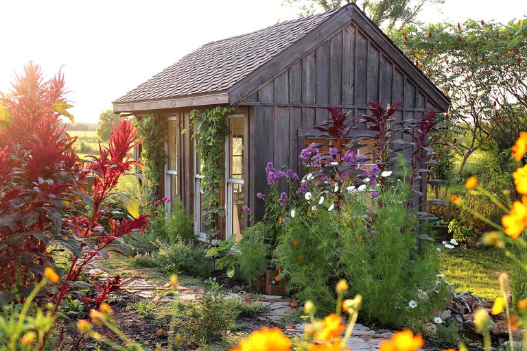 Căsuță de grădină rustică din lemn cu acoperiș în două ape și ferestre mari, înconjurată de o grădină de flori luxuriantă și o potecă de piatră.