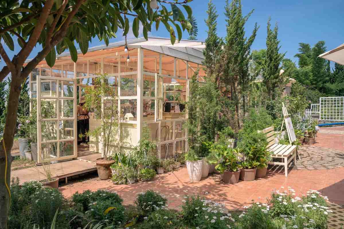 Căsuță de grădină din sticlă și lemn pe o terasă cu plante la ghiveci, copaci înalți, o bancă albă și un șevalet.