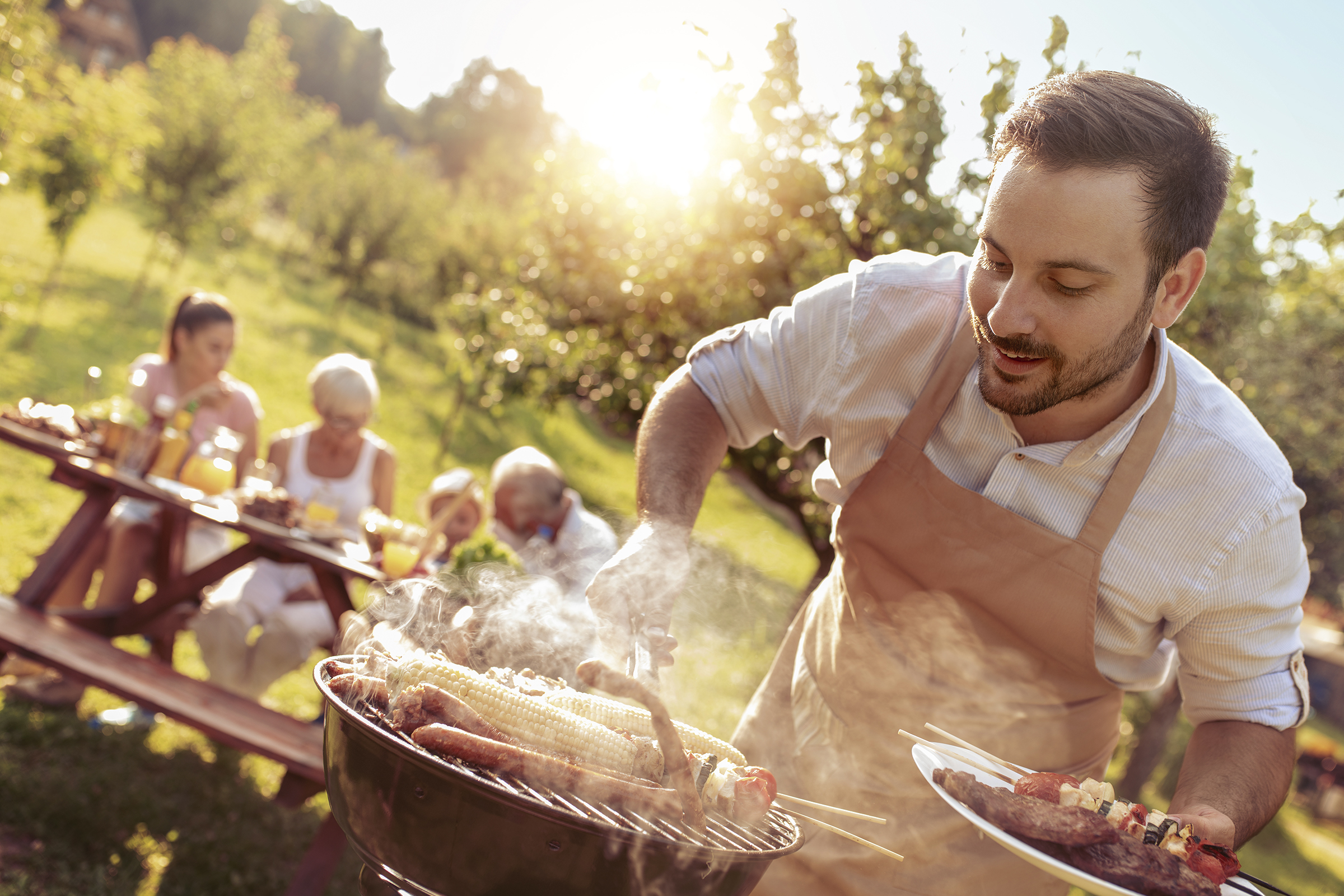 Un bărbat face grătar cu cârnați și porumb pe grătarul cu cărbune din grădină. Un grup de oameni stă la o masă de picnic în fundal.
