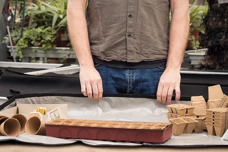 O persoană pregătește o masă cu tăvi de însămânțare, ghivece de plante și pământ pentru grădinărit.