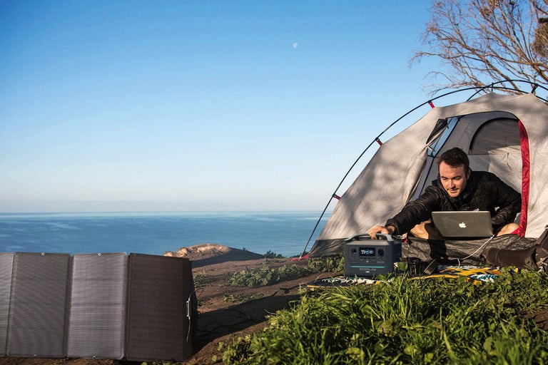 Scenă de camping cu cort, persoană cu laptop, panou solar și stație de alimentare pentru energie electrică independentă de rețea