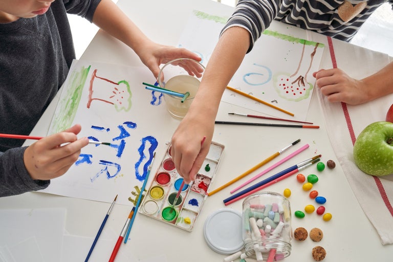 Scenă cu copii pictând cu acuarele, pensule, pahar cu apă, cretă și hârtie, aranjament pe o masă.