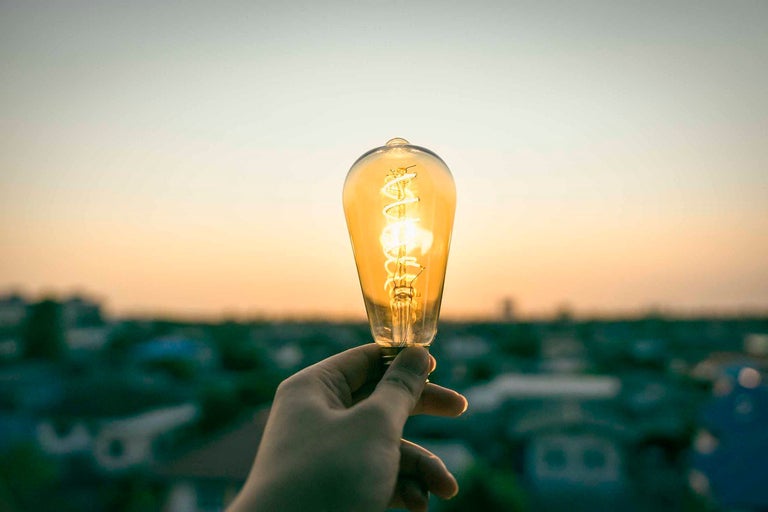 Mână ținând un bec cu filament în fața unui fundal neclar.