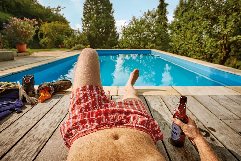 Bărbat se relaxează la piscină cu bere, pantaloni de lucru și pantofi pe terasa de lemn.