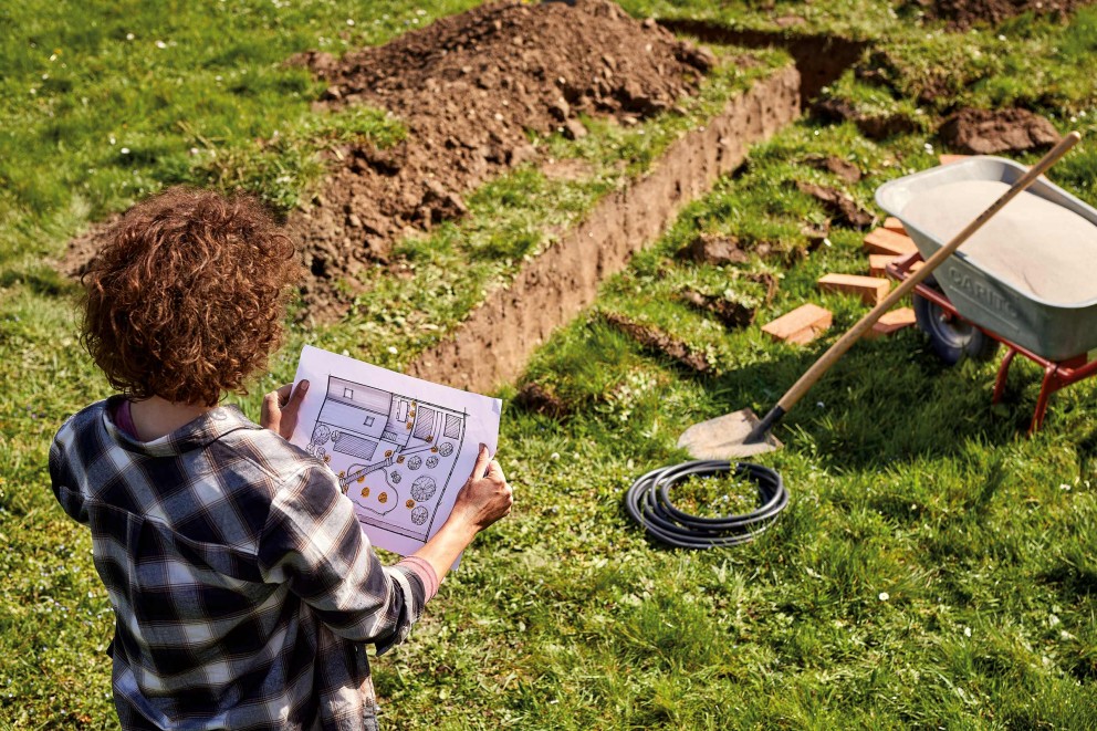 Femeie se uită la planul de construcție în grădină cu lopată, roabă, furtun și șanț săpat