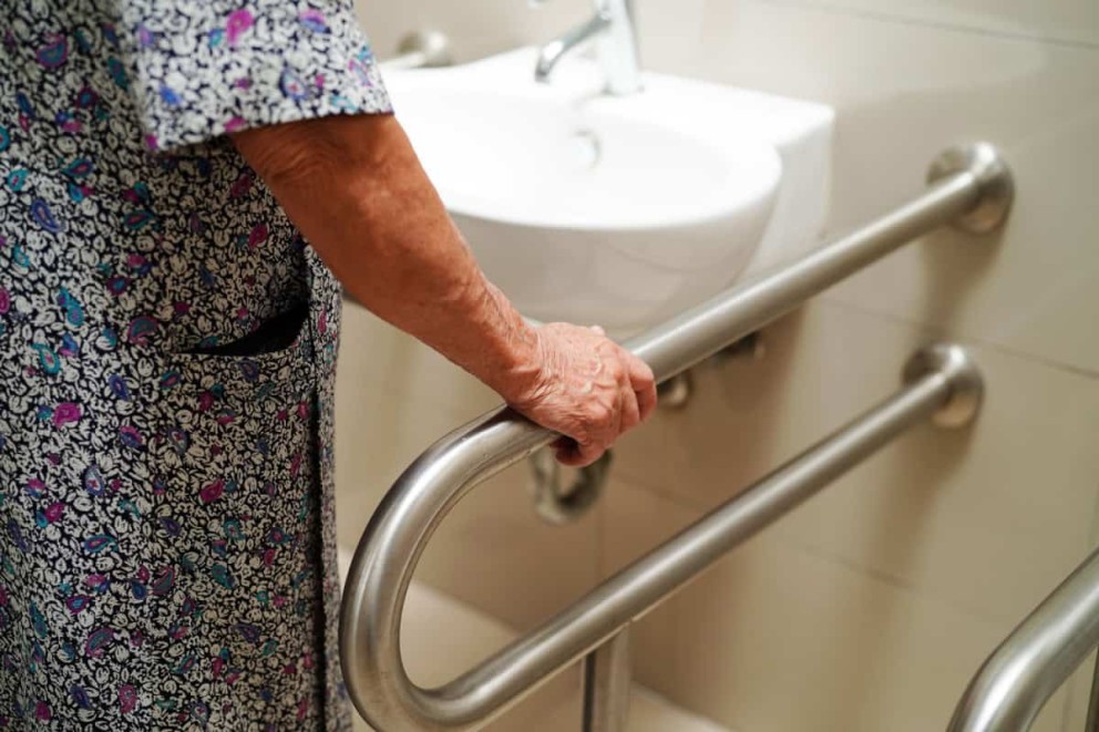 O persoană în vârstă se ține de o bară de sprijin în baie pentru susținere.