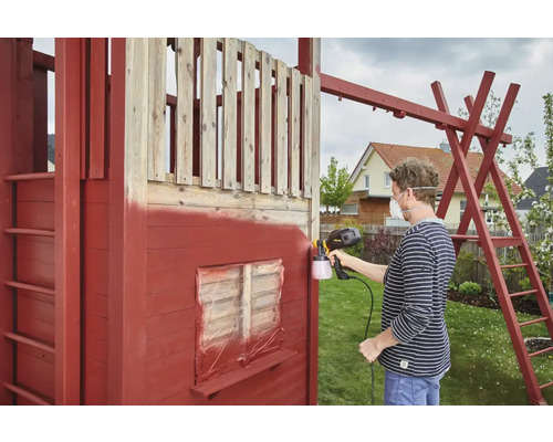 O persoană pulverizează o căsuță de joacă din lemn cu un pistol de pulverizat vopsea