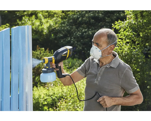 O persoană folosește un pulverizator de vopsea pentru a vopsi un gard din lemn. Persoana poartă o mască de protecție.