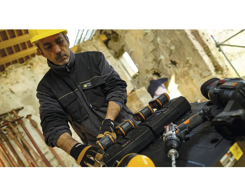 Un muncitor în construcții cu cască de protecție schimbă bateria unei scule electrice pe un șantier.