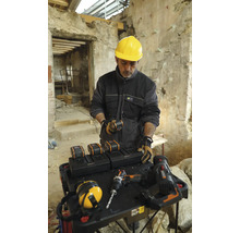 Meșter cu cască de protecție galbenă la o masă cu baterii, încărcătoare și scule într-o clădire.