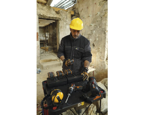 Meșter cu cască de protecție galbenă la o masă cu baterii, încărcătoare și scule într-o clădire.