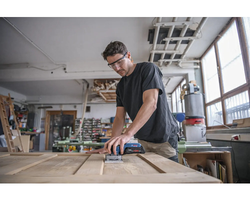 Un bărbat șlefuiește o ușă din lemn cu un șlefuitor oscilant cu acumulator într-un atelier.