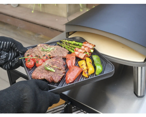 O persoană cu mănuși de cuptor ține o tavă de grătar cu friptură, sparanghel, roșii și ardei grași la grătar în fața unui cuptor de pizza.