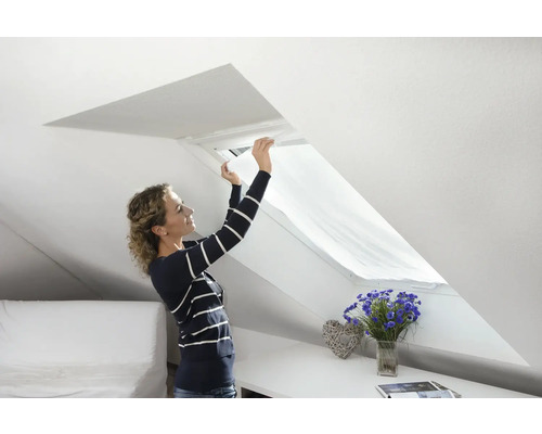 Femeie montând o rolă de protecție solară pe o fereastră de mansardă într-o cameră de mansardă luminoasă.