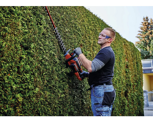 Un bărbat tunde un gard viu cu o foarfecă de gard viu și poartă ochelari de protecție.