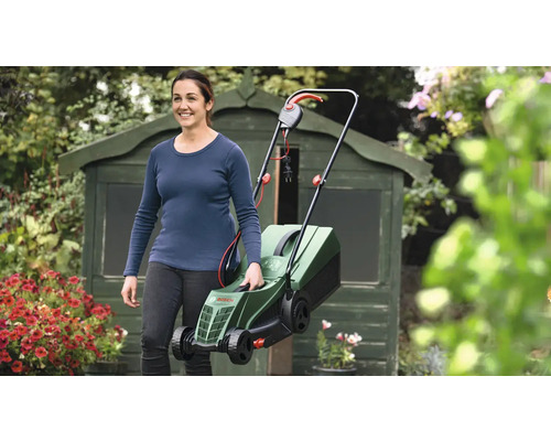 Femeie care poartă o mașină de tuns iarba electrică în grădină în fața unei magazii de scule.