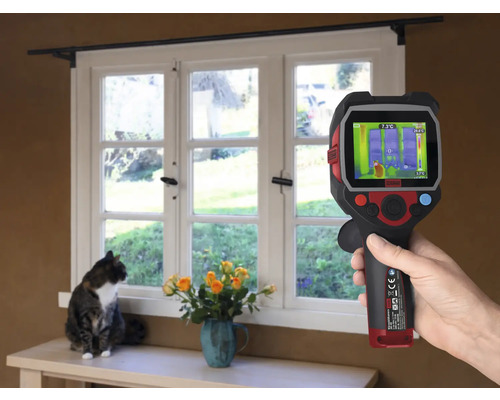 O mână ține o cameră termică în fața unei ferestre, în timp ce o pisică stă pe pervaz.