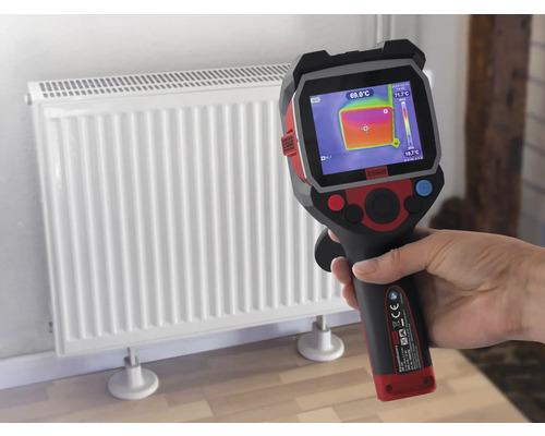 O mână ține o cameră de termoviziune în fața unui radiator pentru a măsura temperatura.