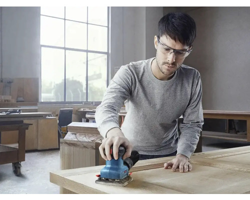 Un bărbat șlefuiește lemn cu o șlefuitoare vibratoare într-un atelier.