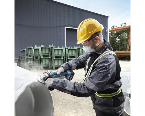 Un bărbat cu cască de protecție, ochelari de protecție și mască de protecție șlefuiește o piesă de lucru cu o polizor unghiular cu acumulator.