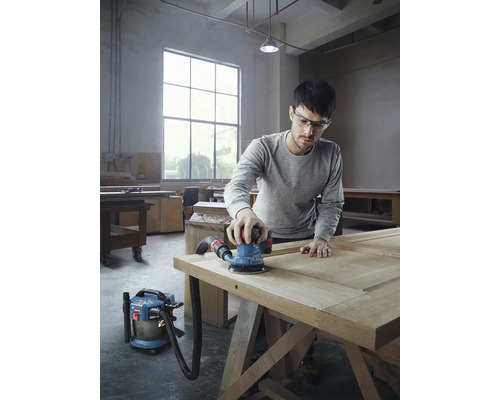 Un bărbat șlefuiește o ușă din lemn cu o șlefuitoare excentrică și un aspirator într-un atelier.