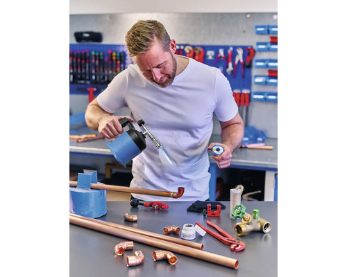 Un bărbat sudează țevi de cupru cu un arzător, fludor și diverse instrumente pe un banc de lucru.