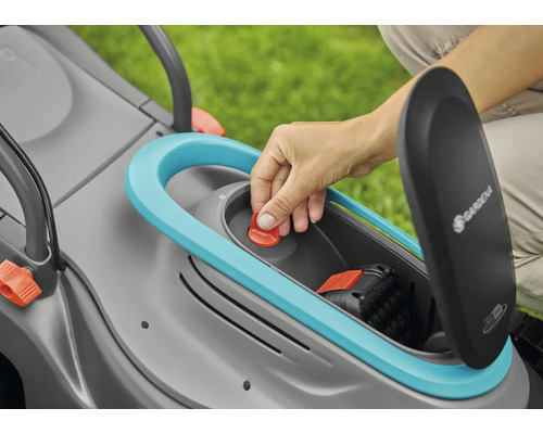 O mână introduce un buton roșu într-o mașină de tuns iarba cu un compartiment pentru baterii deschis. Logo-ul Gardena este vizibil.