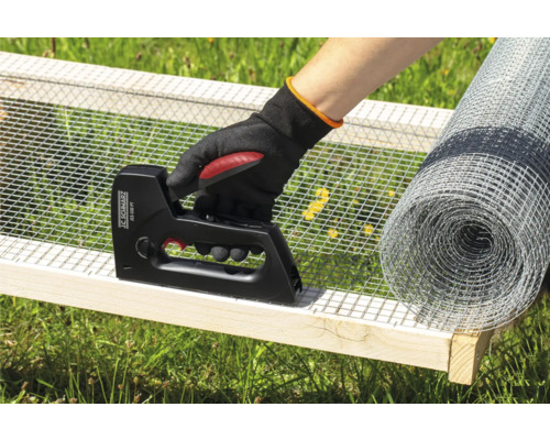 O persoană fixează plasă de sârmă cu un capsator pe un cadru de lemn, cu o rolă de plasă de sârmă în fundal.