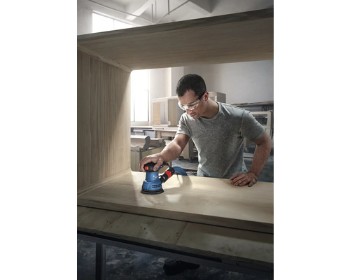 Un bărbat șlefuiește o piesă de mobilier din lemn cu un șlefuitor excentric marca Bosch.