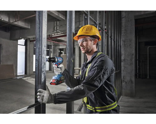 Un muncitor cu cască de protecție și ochelari de protecție folosește o șurubelniță cu acumulator pe un șantier.