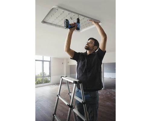 Un bărbat fixează o plafonieră cu LED cu o șurubelniță fără fir într-o cameră, stând pe o scară.