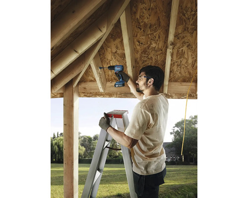 Un meșter forează o gaură cu o șurubelniță de găurit cu acumulator într-o grindă de lemn sub acoperișul unei construcții brute, în timp ce stă pe o scară.