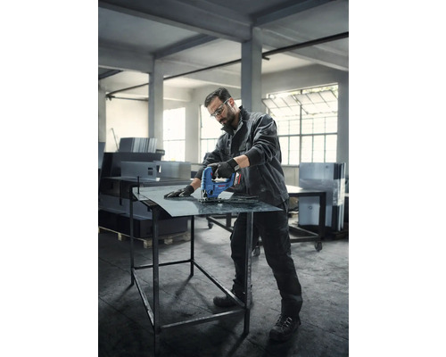 Un bărbat cu ochelari de protecție și mănuși taie o foaie de metal cu un ferăstrău vertical pe o masă de lucru.